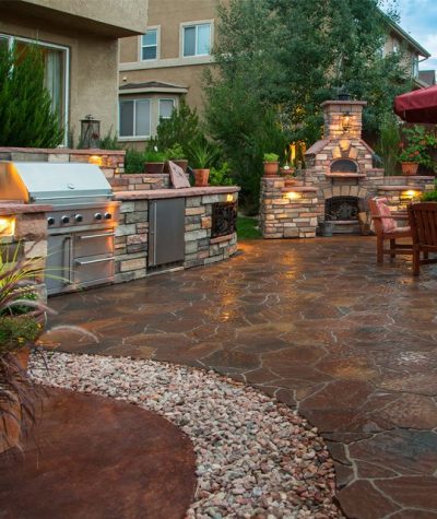 Paver patio with a fire pit, outdoor kitchen, pizza oven and lighting at dusk.